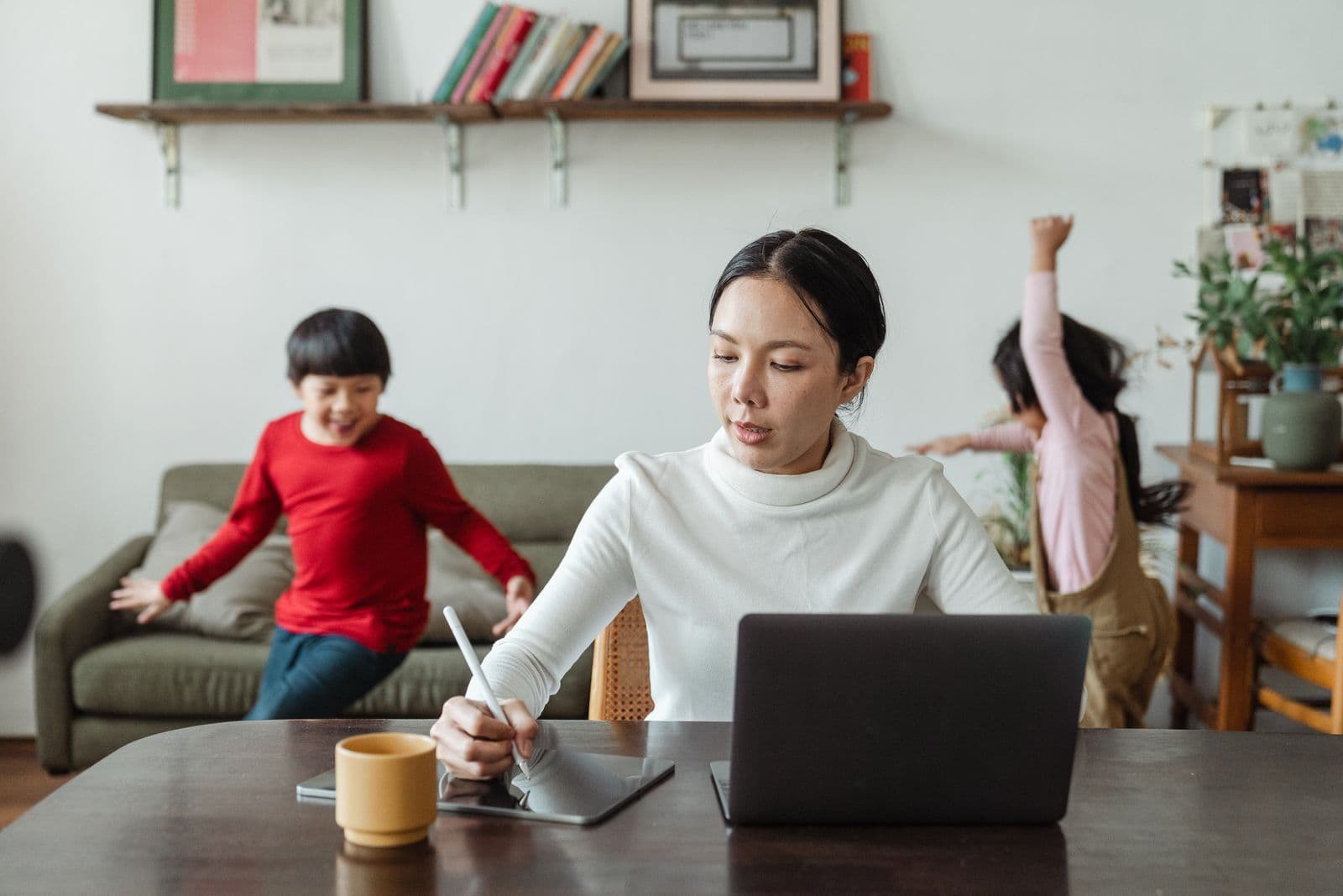 East Asian mother working from home while her children play energetically behind her