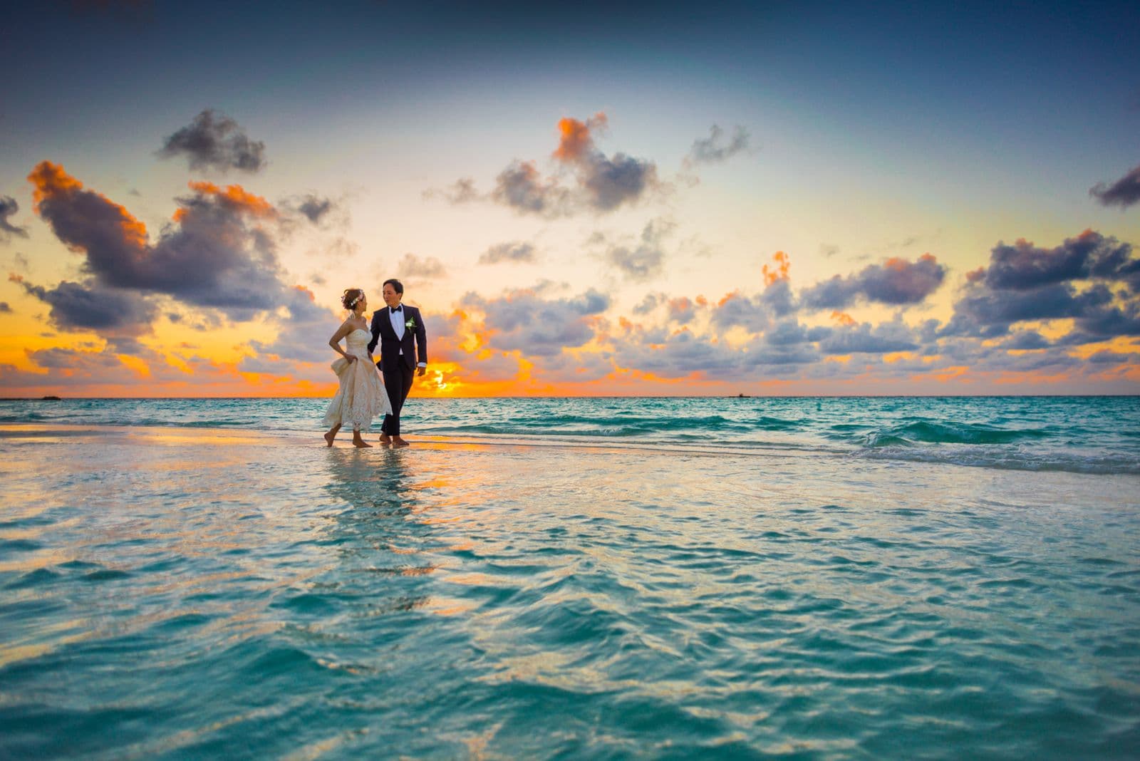 East Asian bride and groom walking hand in hand along a beach at sunset