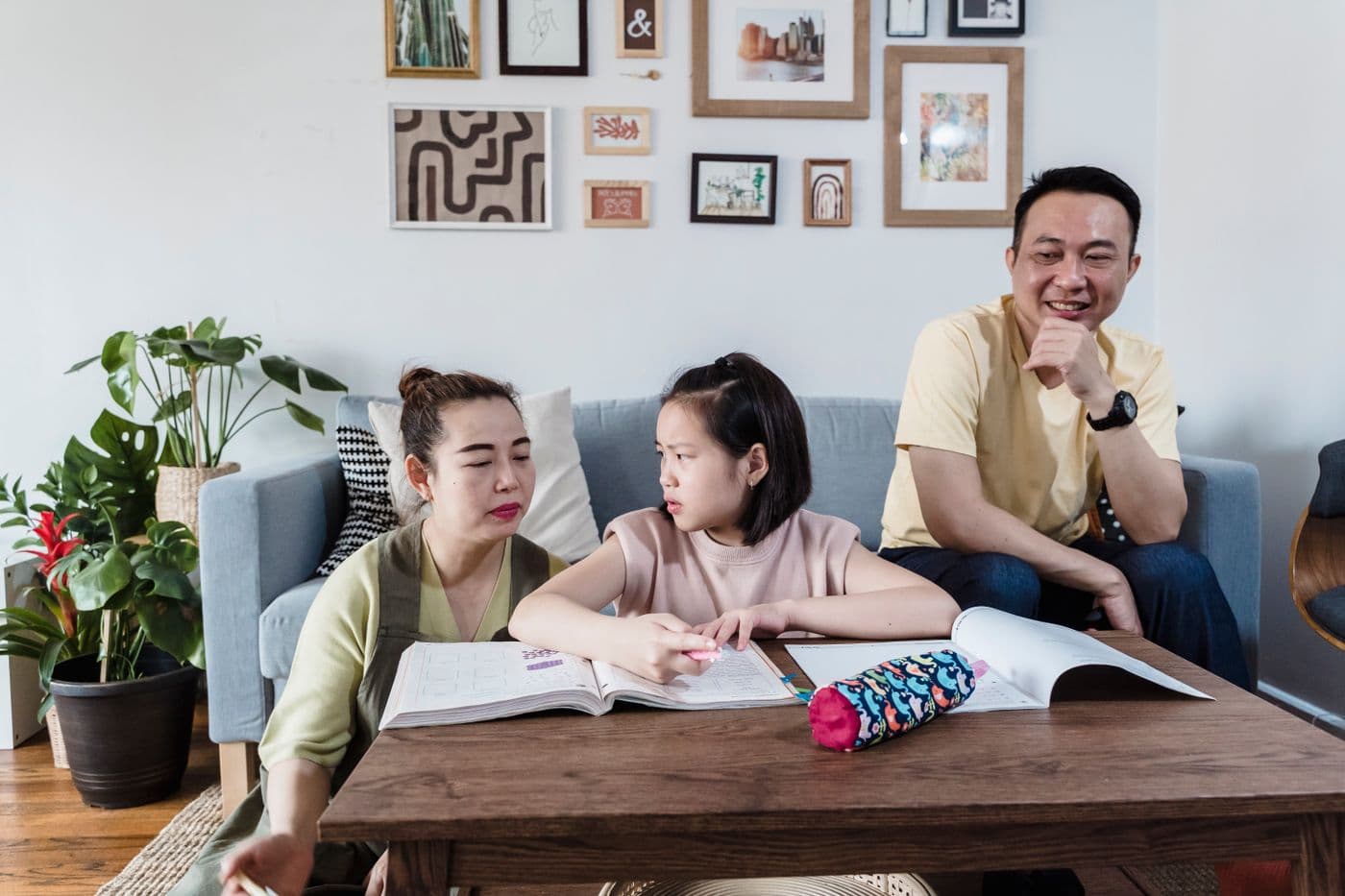 East Asian mother helping her daughter with homework at the coffee table while father watches from the sofa