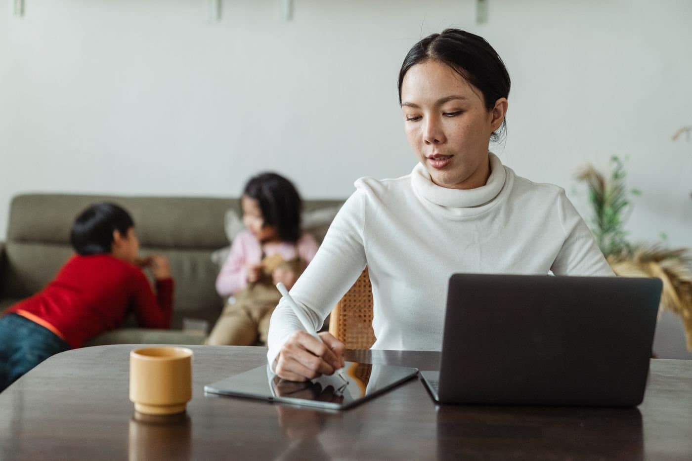 East Asian mother working at home while her children play on the sofa behind her