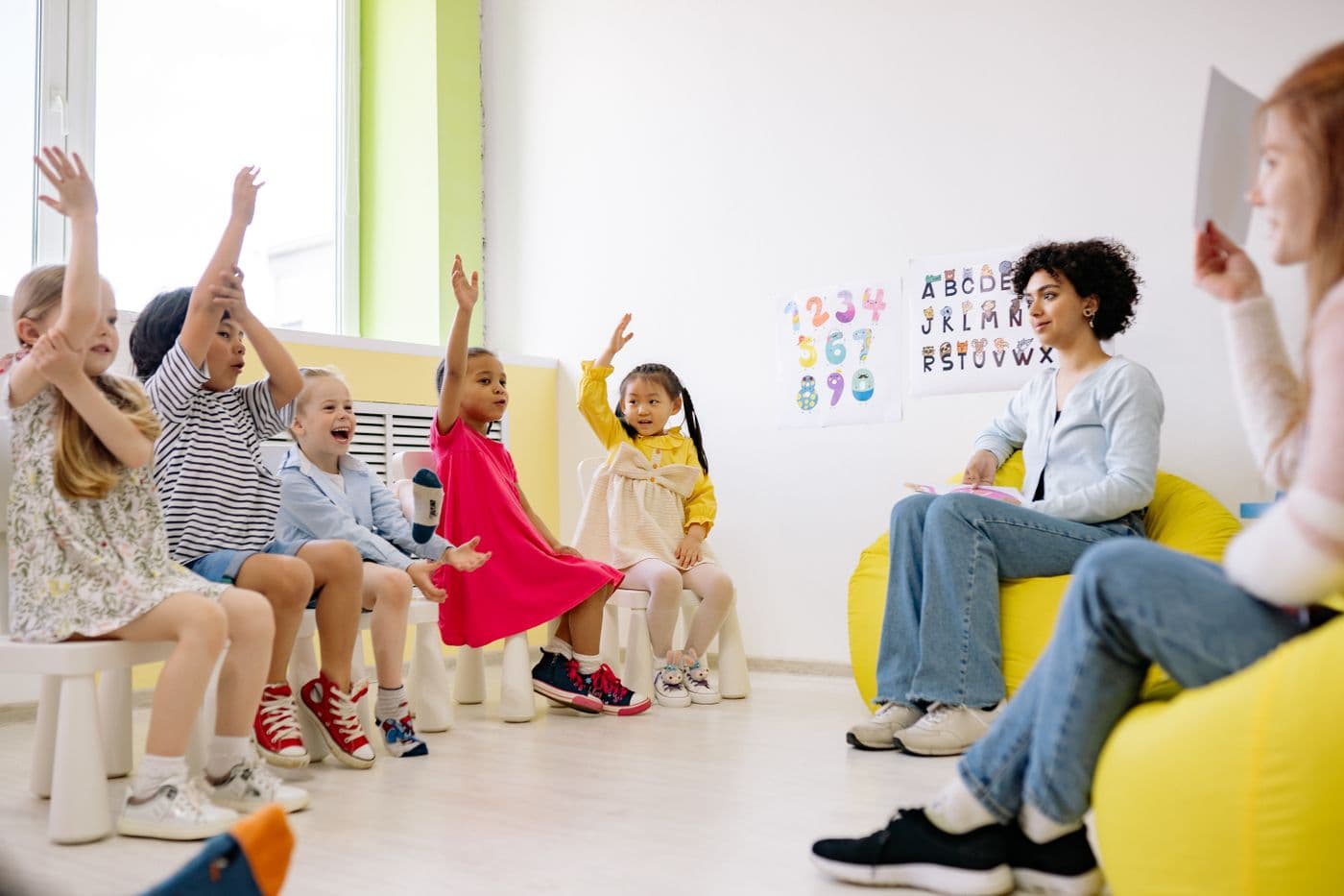 Diverse preschool children raising hands in a bright classroom with a teacher