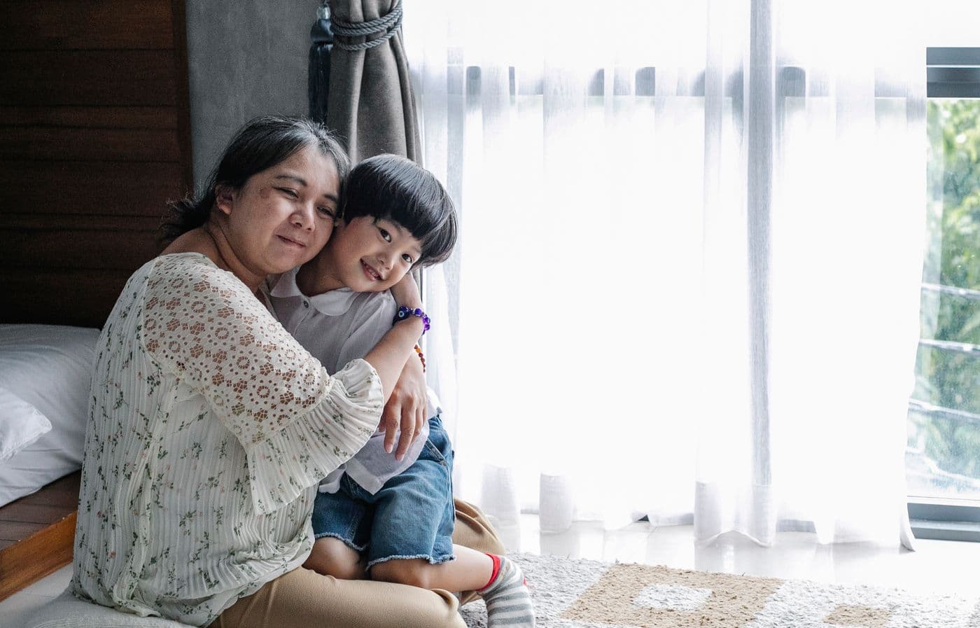 East Asian grandmother embracing her young grandson in a sunlit bedroom during a visit