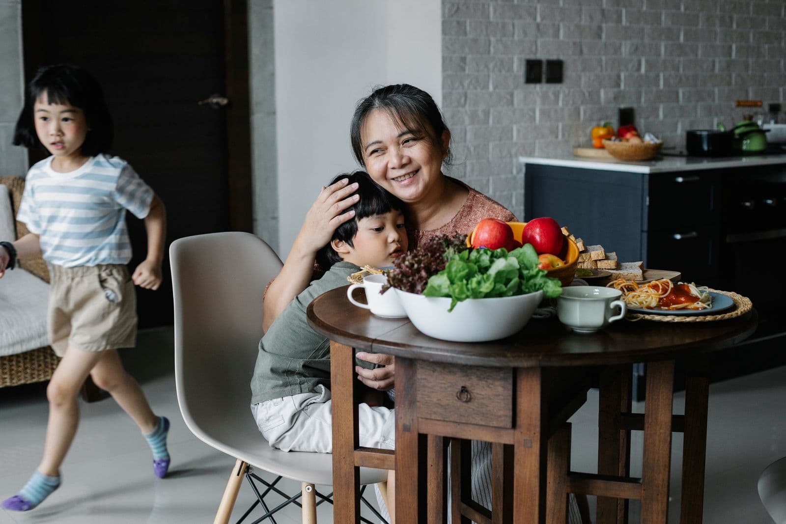 Grandmother embracing one grandchild at the table while another child runs past