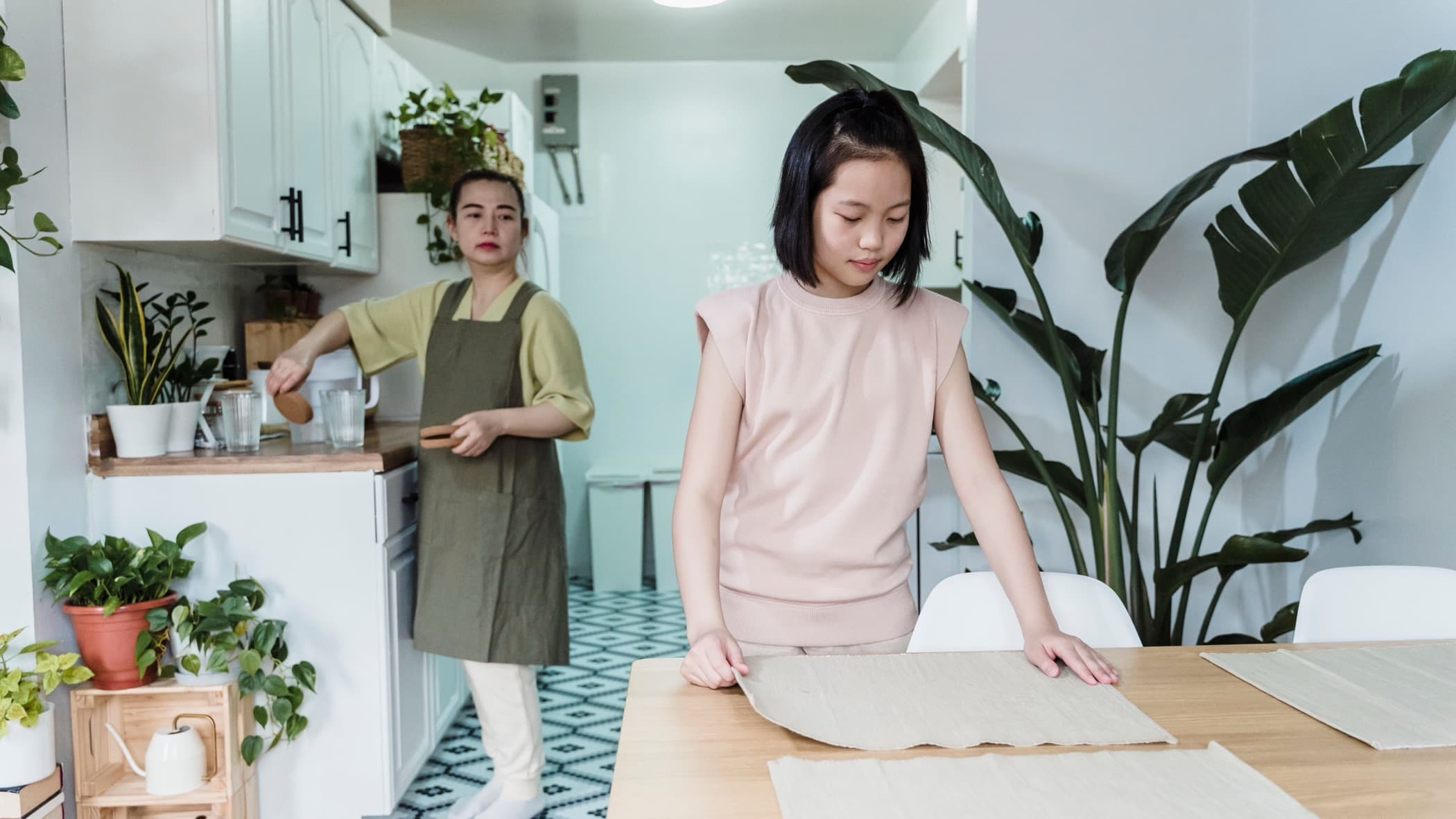 An East Asian woman sets the dining table while her mother works at the kitchen counter