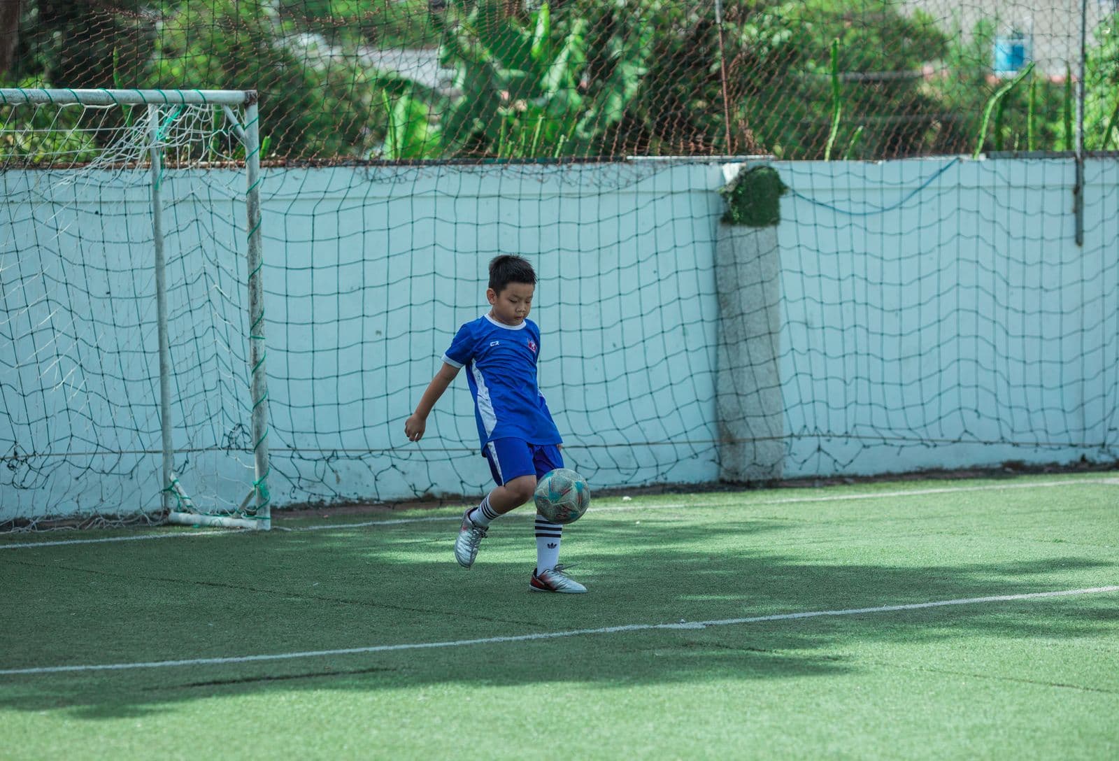 East Asian boy in a blue soccer kit practicing ball control on an outdoor turf field