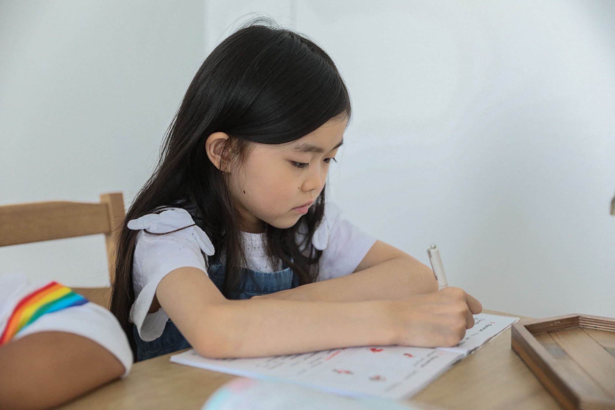 East Asian child writing in an open workbook with red markings at a light wood table