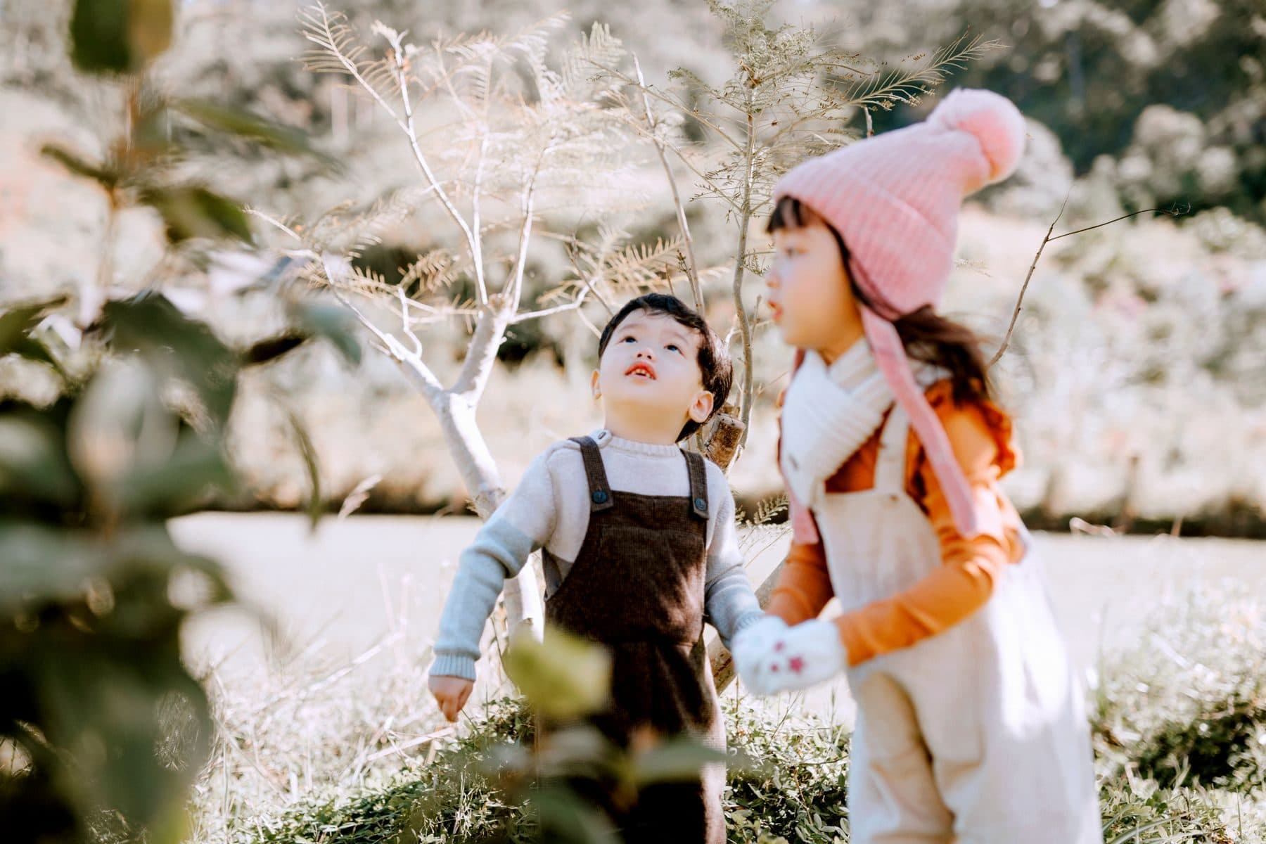 Two East Asian children wandering outdoors among grass and thin trees on an ordinary afternoon