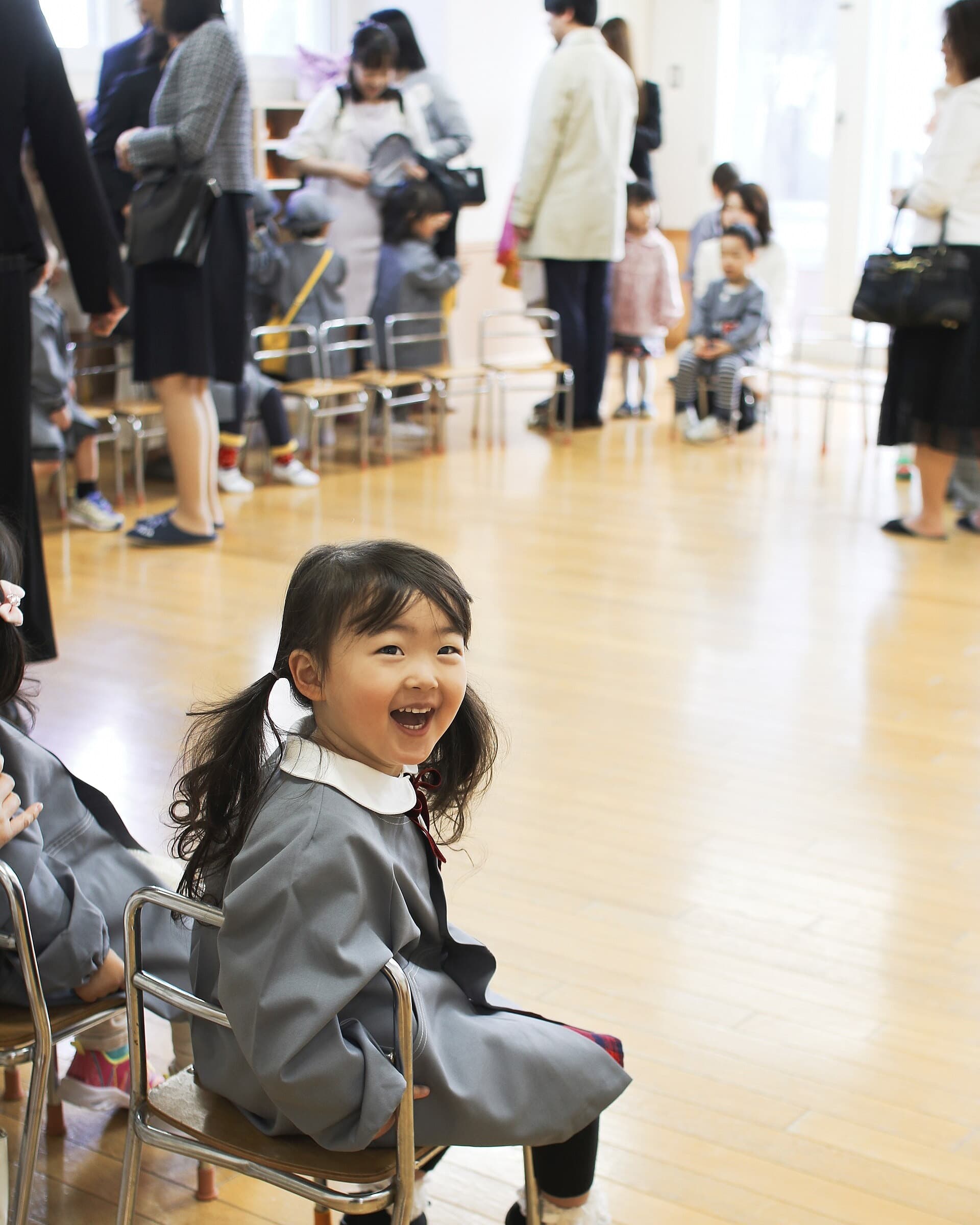 East Asian preschooler laughing in a kindergarten classroom while other children and adults move through the room behind her