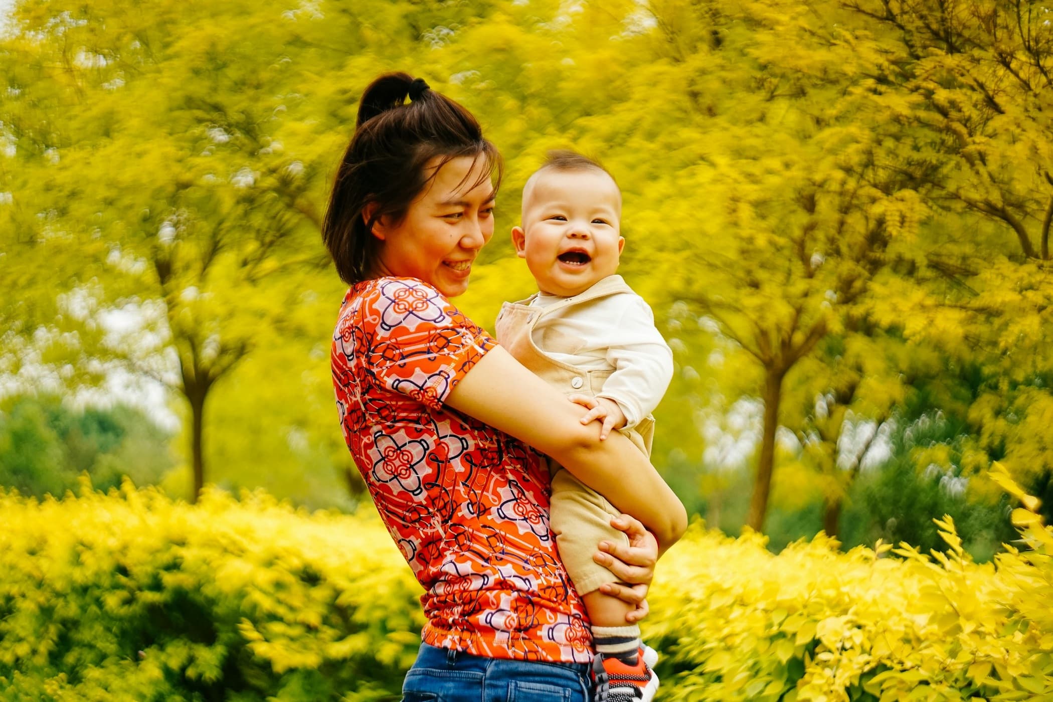 East Asian mother smiling as she holds her baby outdoors among yellow-green trees