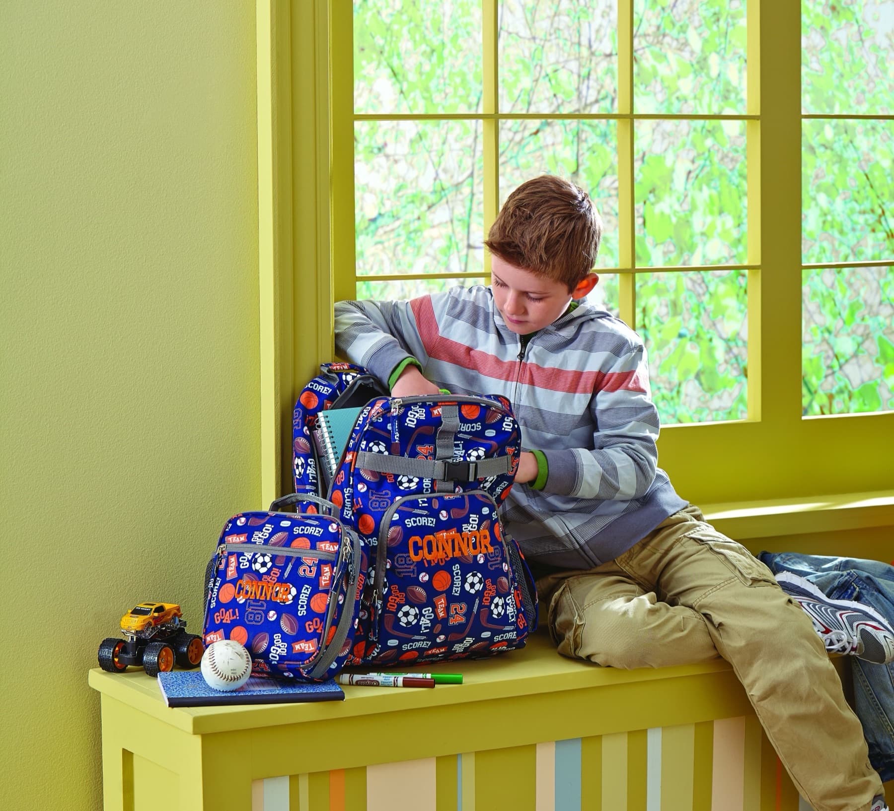 School-age boy sitting on a window bench with a sports backpack, baseball, notebook, and toys nearby