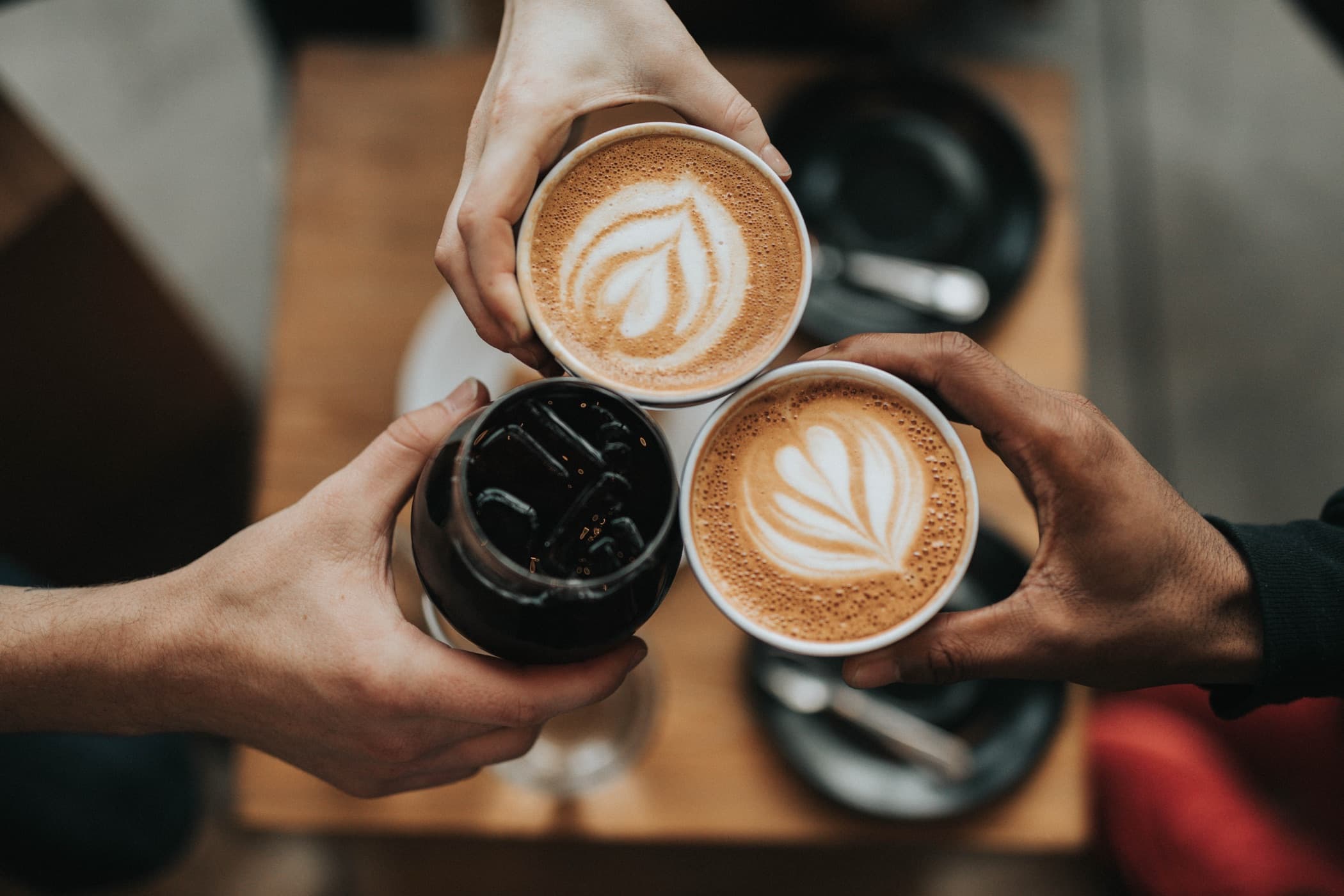 Three people toasting coffee cups together over a wooden cafe table