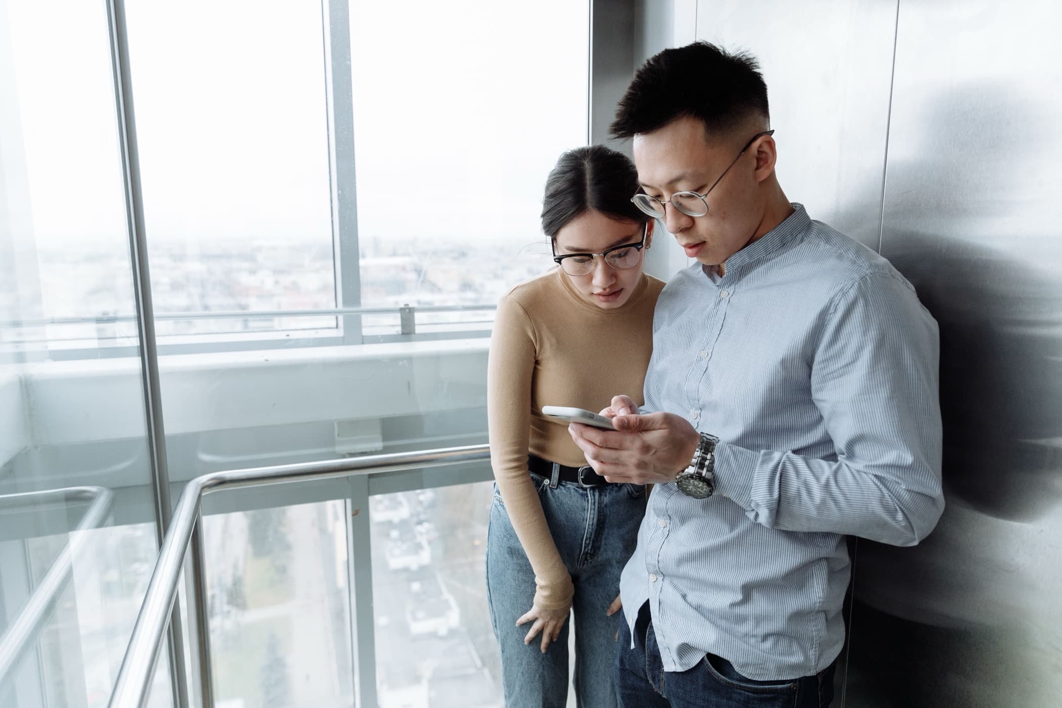 An East Asian man and woman leaning together over a smartphone in a glass elevator with a city view