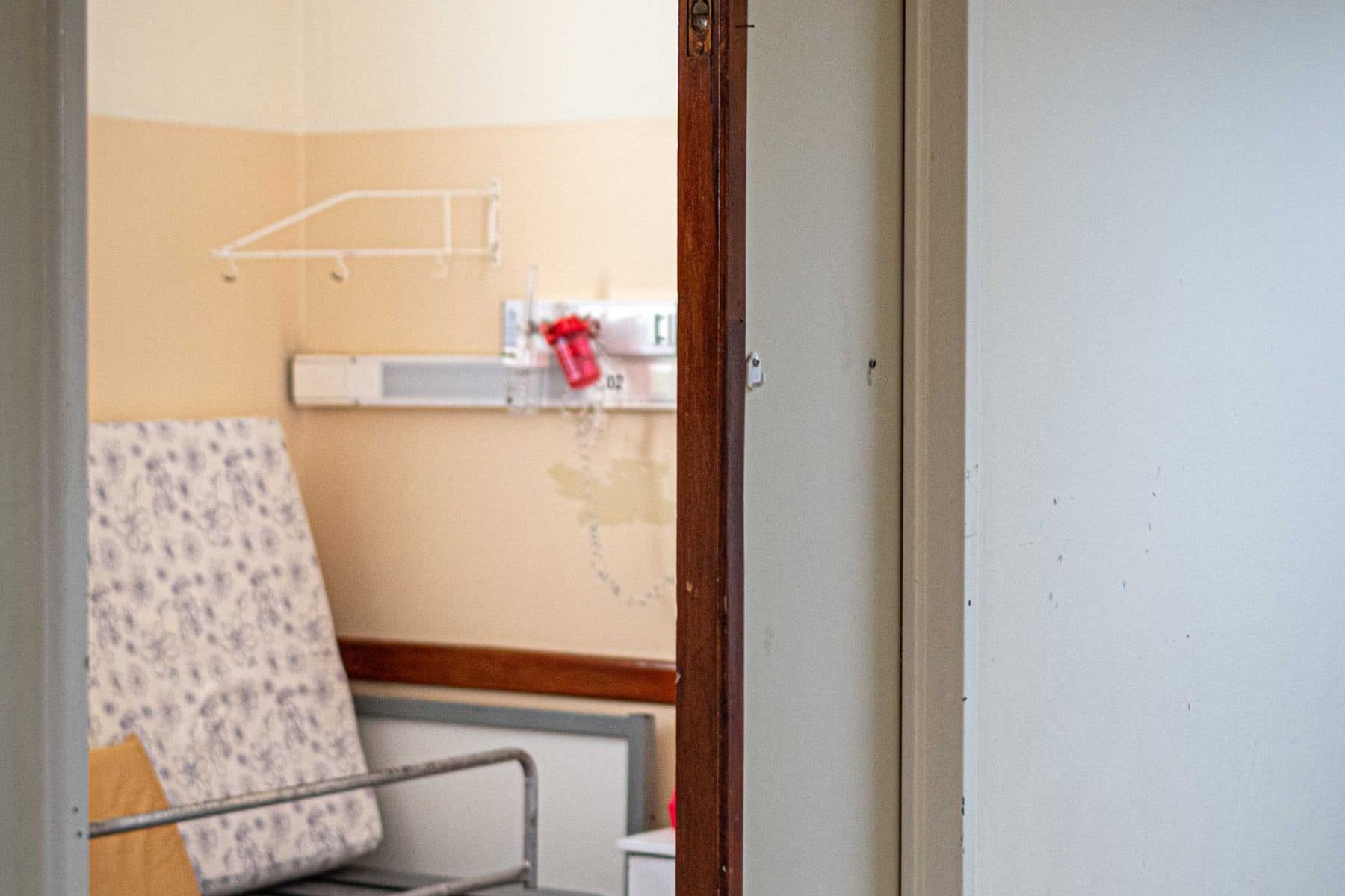 Empty hospital room seen through an open doorway, with a bed frame and medical wall fixtures