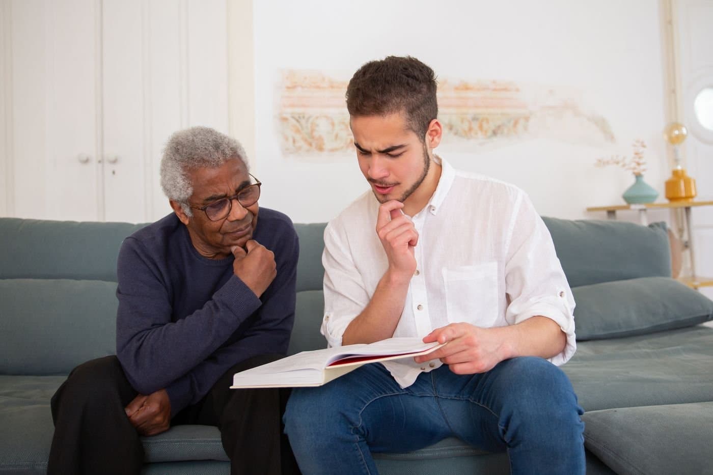 Young adult man and older man sitting side by side on a living-room sofa, reading a book together