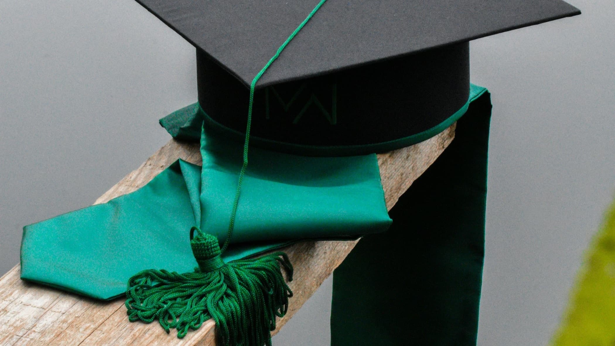 Black graduation cap with a green tassel and stole resting on a wooden rail