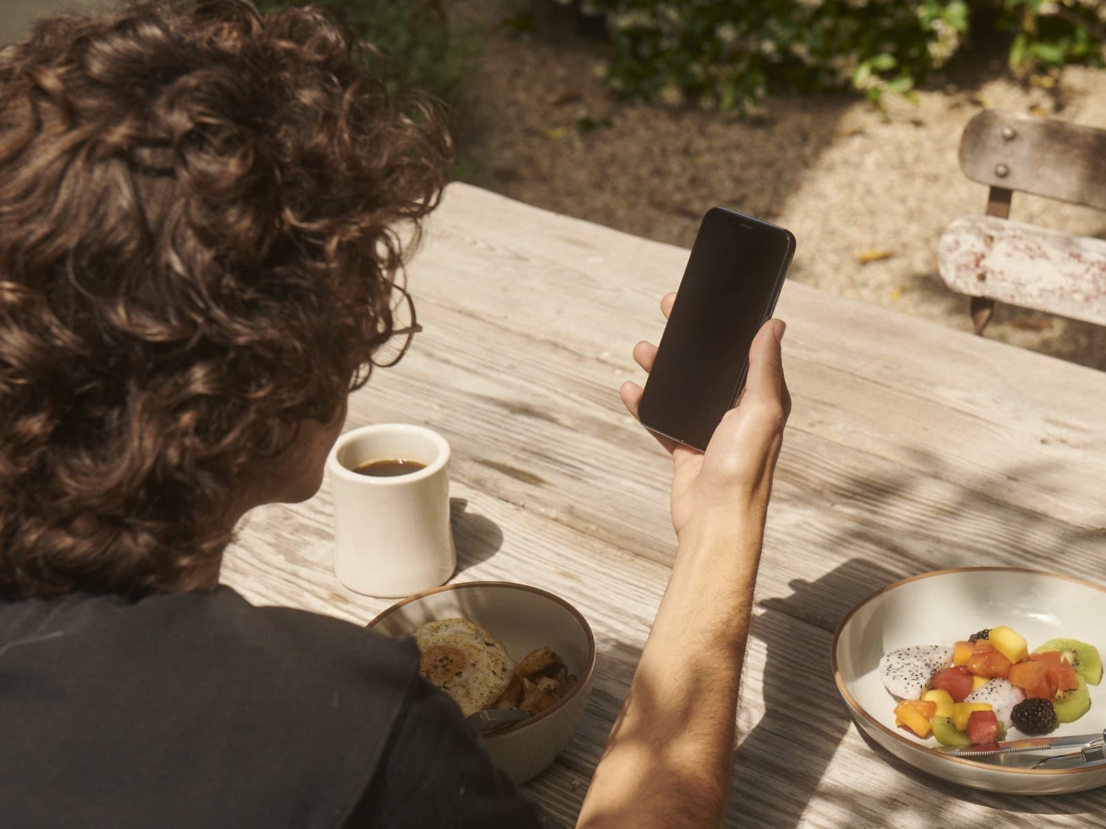 Person checking a phone at an outdoor breakfast table with coffee and fruit