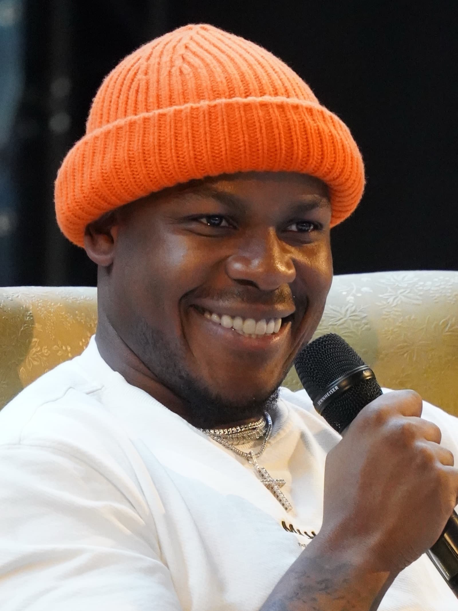 John Boyega smiling in an orange beanie and white T-shirt, holding a microphone at Comic Con Brussels