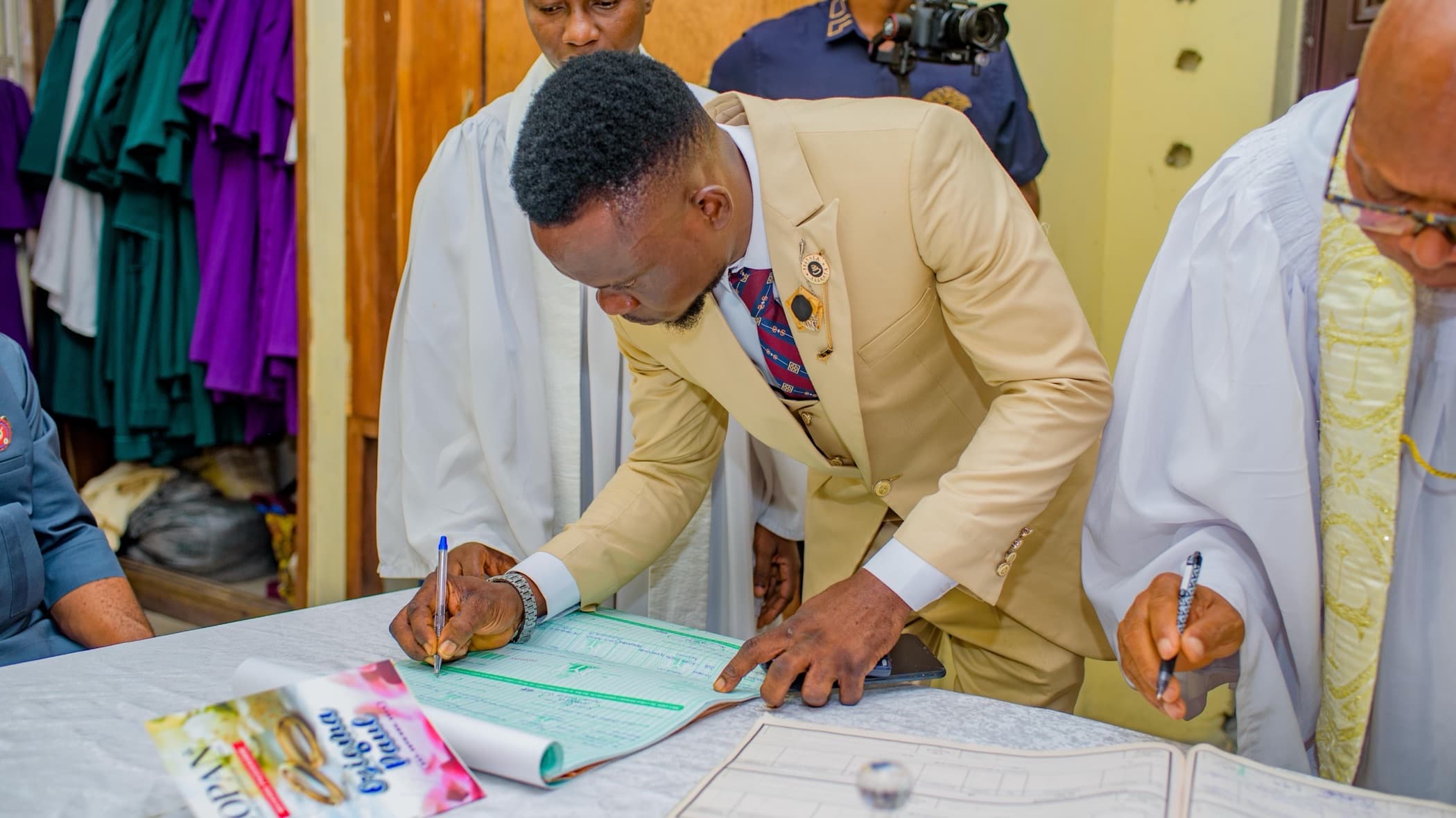 A groom in a tan suit signs a marriage register at a table while clergy stand beside him