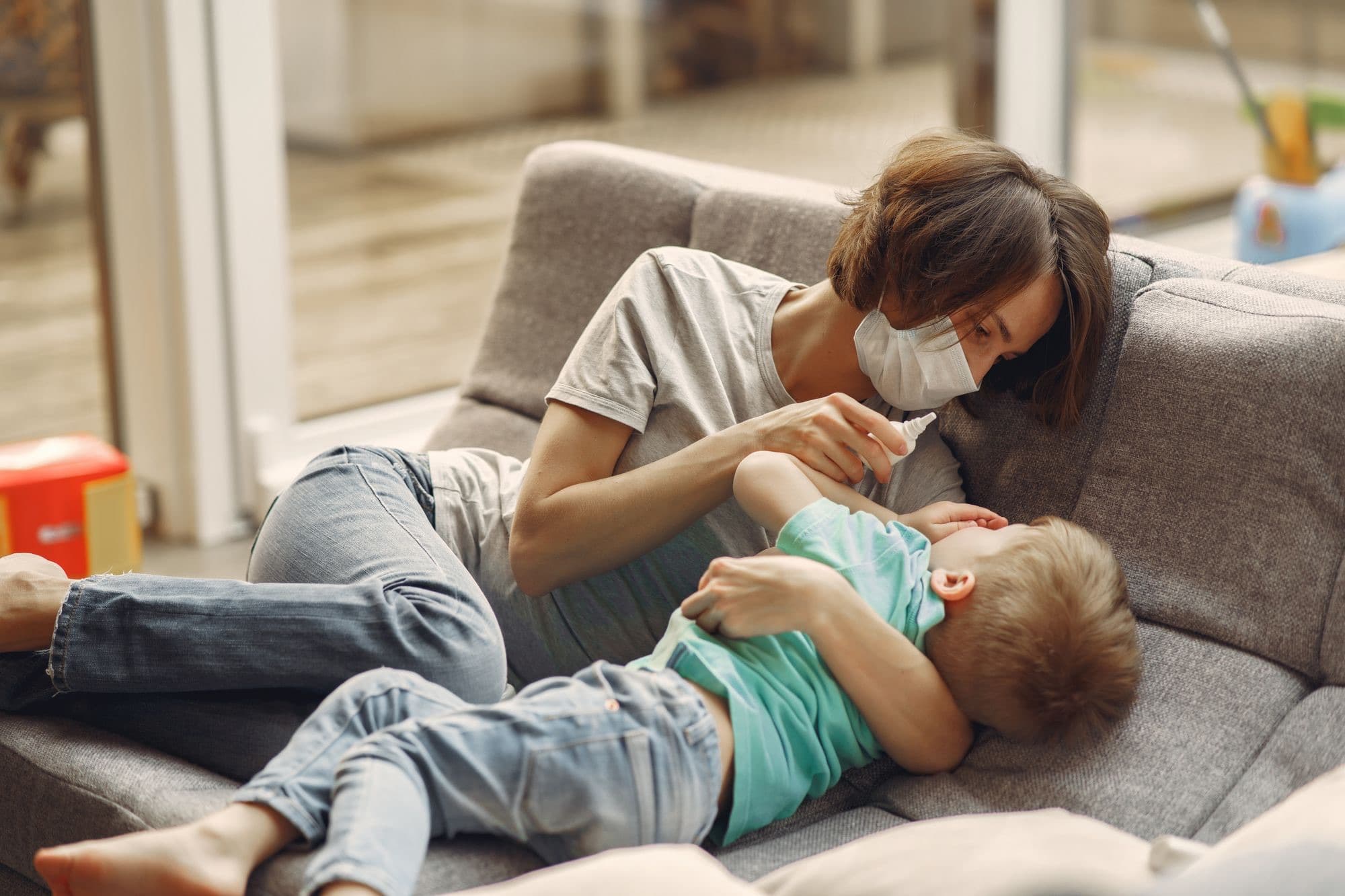 A parent in a face mask gives nasal spray to a child lying on a gray couch