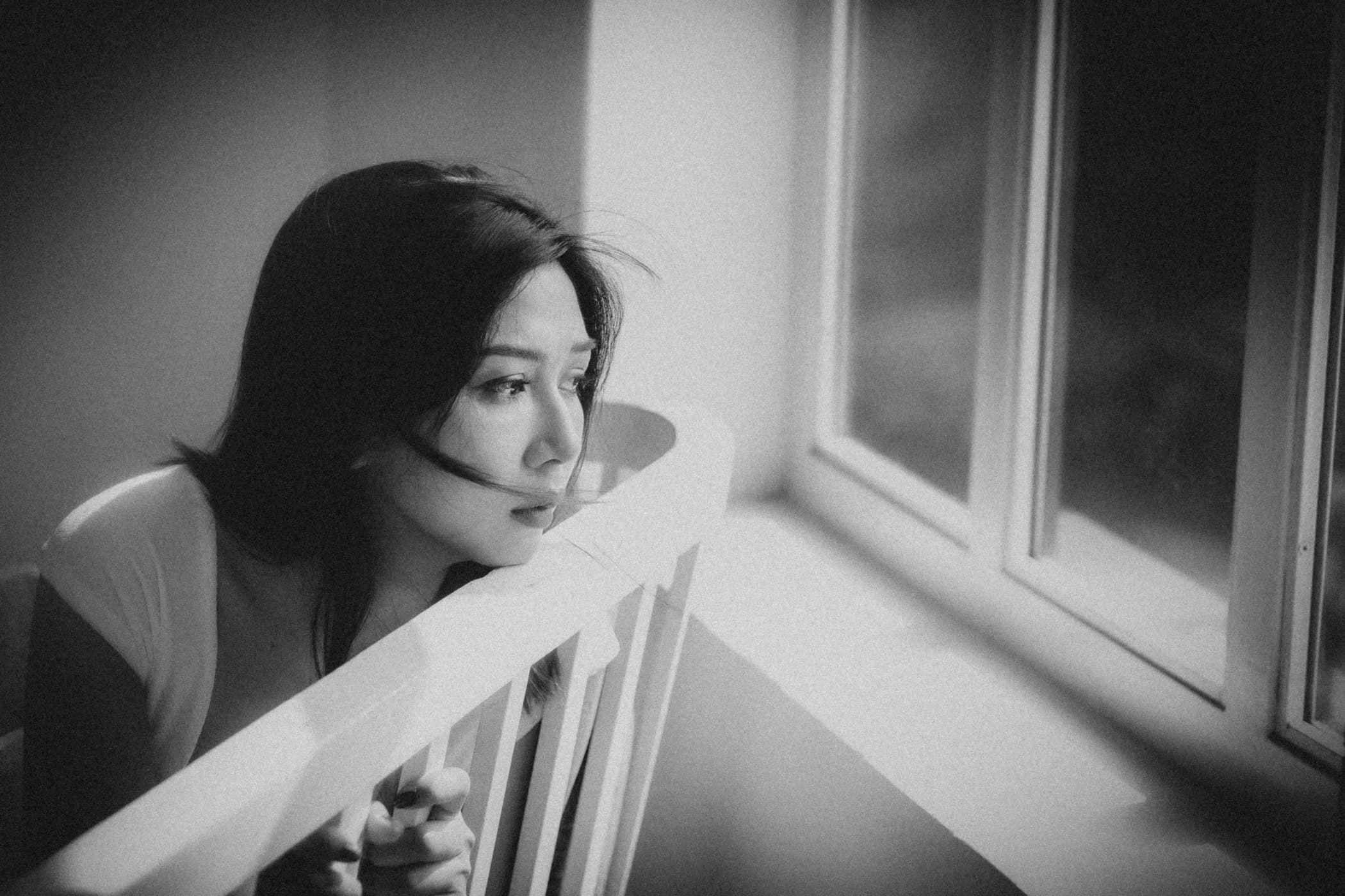A young East Asian woman resting her chin on a railing while looking out a bright window