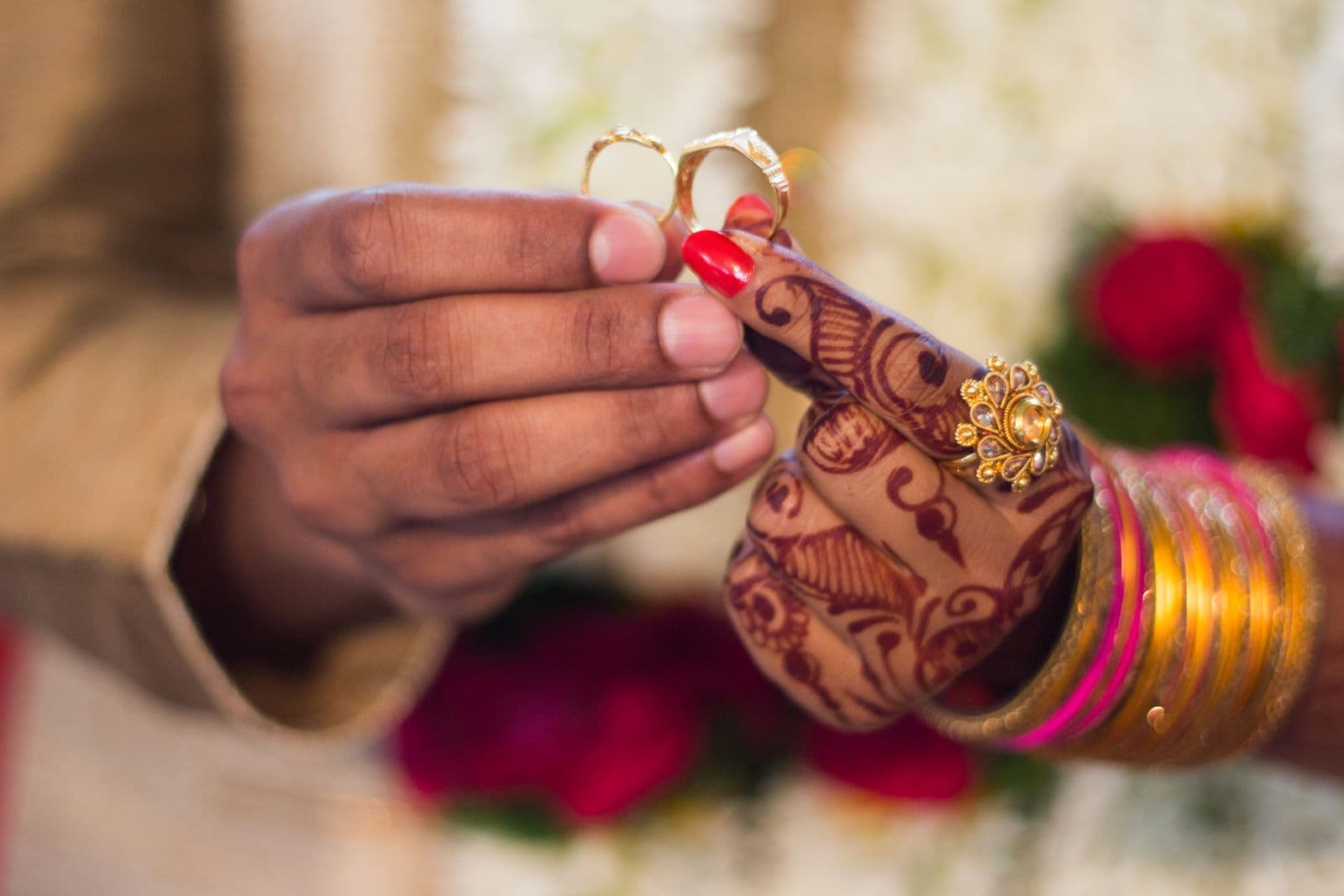 Couple holding gold wedding bands during a South Asian ceremony with henna and bangles