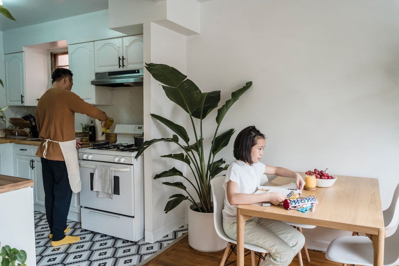 East Asian father cooking in the kitchen while his daughter studies at the table